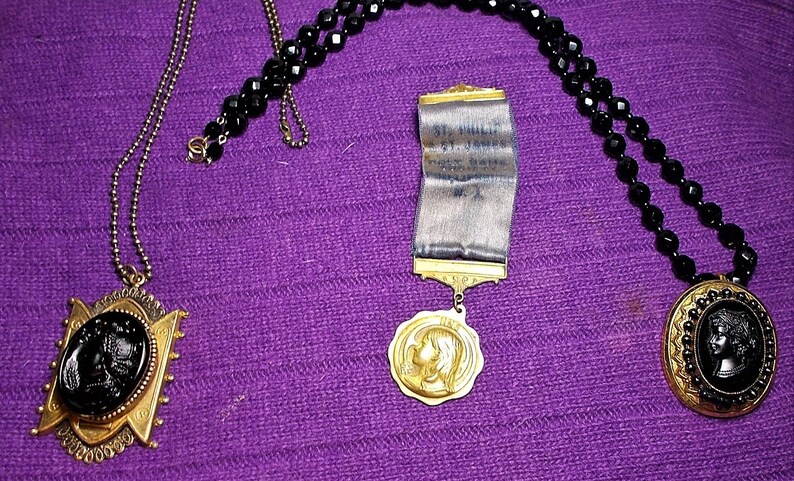 May include: Three necklaces with black beads and gold pendants. One pendant features a black cameo of a woman's profile, another has a gold medal with a woman's profile, and the third has a black cameo of a woman's profile in a gold frame with a ribbon that reads "St. Hilda's Guild, 1900, New York, N.Y."