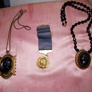 May include: Three necklaces with black cameo pendants. The first necklace has a gold chain and a cameo pendant with a woman's profile. The second necklace has a blue ribbon and a gold cameo pendant with a woman's profile. The third necklace has black beads and a gold cameo pendant with a woman's profile.