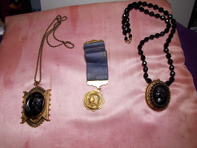 May include: Three necklaces with black cameo pendants. The first necklace has a gold chain and a cameo pendant with a woman's profile. The second necklace has a blue ribbon and a gold cameo pendant with a woman's profile. The third necklace has black beads and a gold cameo pendant with a woman's profile.