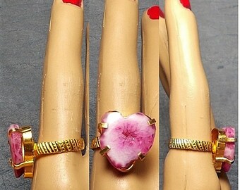 Pode incluir: Um anel de ouro com uma pedra de ágata rosa em forma de coração. O anel está no dedo de um manequim.