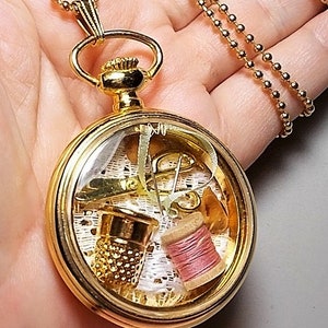 May include: A gold-tone necklace with a round locket pendant. The locket is open and contains miniature sewing supplies, including a thimble, spool of thread, scissors, and pins.