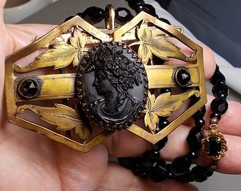 May include: A gold-toned, ornate, openwork pendant with a black cameo featuring a woman's profile. The pendant is attached to a black beaded necklace with a smaller black cameo pendant.