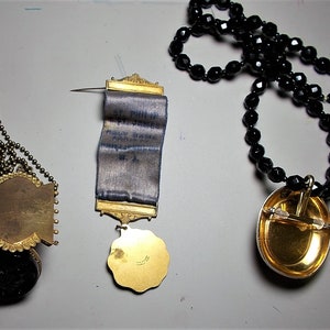 May include: Three gold-toned pendants with different designs. The first is a round pendant with a chain, the second is a rectangular pendant with a blue ribbon, and the third is an oval pendant with a black beaded chain.