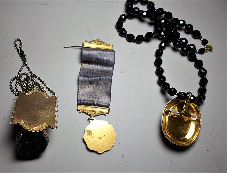 May include: Three gold-toned pendants with different designs. The first is a round pendant with a chain, the second is a rectangular pendant with a blue ribbon, and the third is an oval pendant with a black beaded chain.