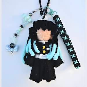 Op de afbeelding: Een handgemaakte pluche pop van een personage met zwart haar en lichtblauwe accenten. De pop heeft een zwarte outfit met gele knopen en een witte riem. Het is bevestigd aan een zwart koord, een lichtblauwe bedel en een zwarte en lichtblauwe band.