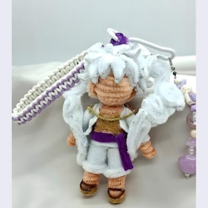 Może przedstawiać: Ręcznie robiona pluszowa lalka z białymi włosami i ubraniami, fioletowym paskiem i brązowymi butami. Lalka ma biały pasek z fioletowo-białym wzorem. Do lalki przymocowany jest mały urok.