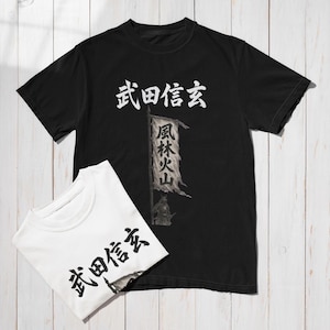 Puede incluir: Camiseta negra con caracteres japoneses blancos y una ilustración de bandera. Una camiseta blanca doblada con el mismo texto está al lado. La ilustración de la bandera tiene caracteres japoneses y una figura de guerrero.