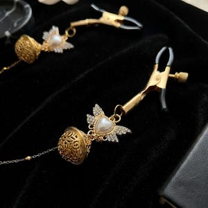May include: Two gold-toned metal clamps with black rubber-coated tips, each featuring a decorative charm. The charms consist of a heart-shaped faux pearl with crystal-encrusted wings attached to a filigree gold-toned bell, connected by a thin chain.