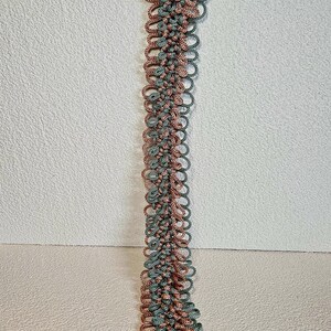 May include: A long, handmade macrame plant hanger in shades of dusty rose and teal green. The hanger is suspended from a metal ring and features a detailed, looped design. The bottom has a tassel.