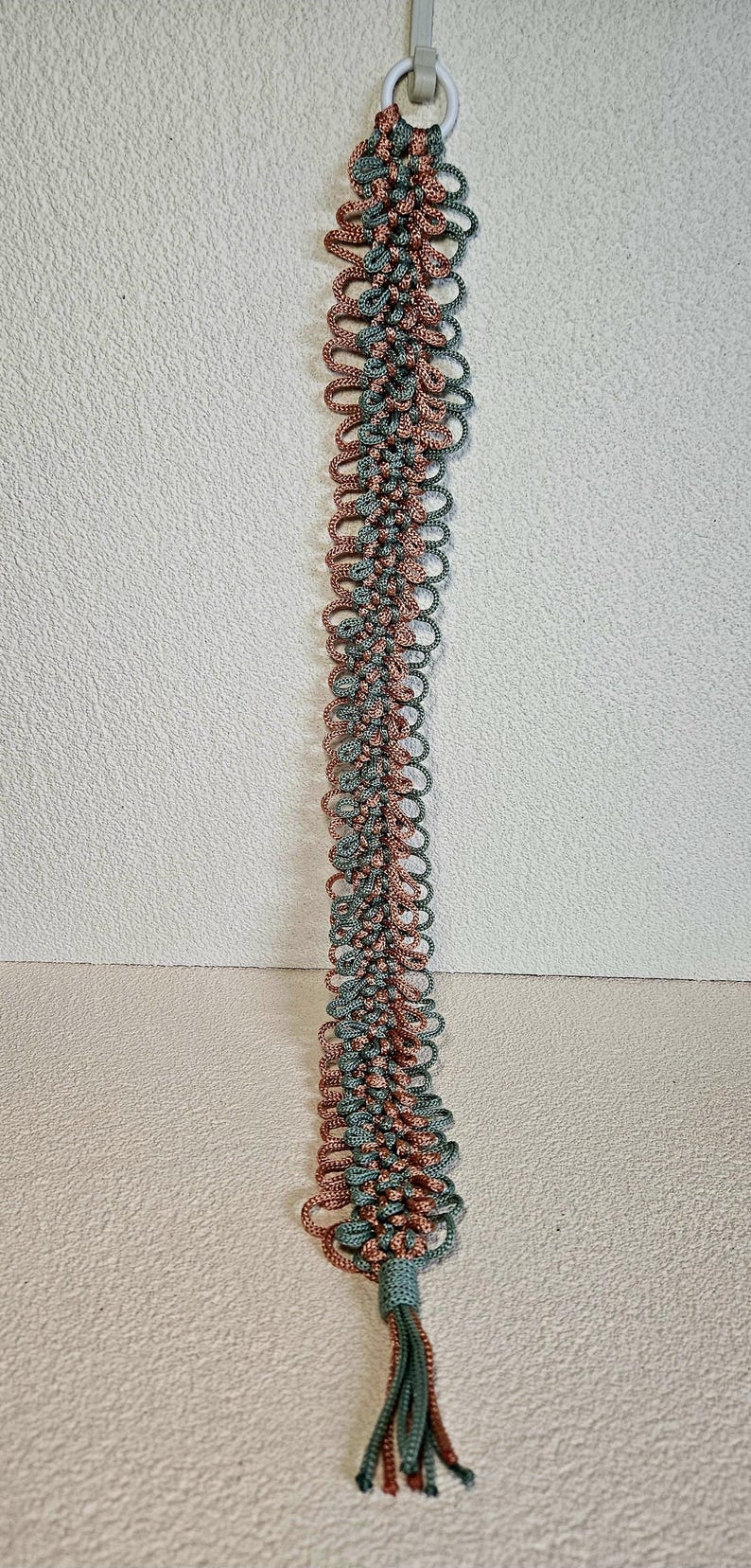 May include: A long, handmade macrame plant hanger in shades of dusty rose and teal green. The hanger is suspended from a metal ring and features a detailed, looped design. The bottom has a tassel.