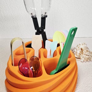 May include: An orange, spiral-shaped organizer filled with various kitchen utensils. Includes a clear plastic scoop, wooden and plastic spoons, and a green spatula. The organizer is on a white surface.