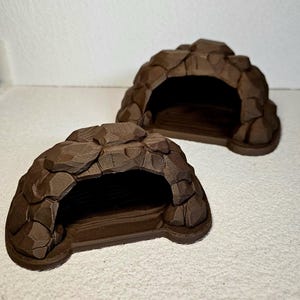 May include: Two brown, cave-shaped pet habitats. Each has a rounded, rocky exterior with an arched opening. The interiors are dark, suggesting a cozy space. The structures are designed to resemble natural rock formations, providing a secure and visually appealing shelter.