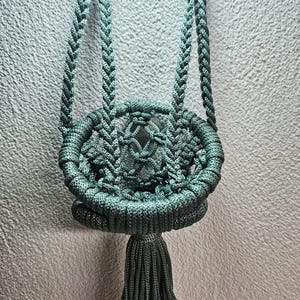 May include: A teal macrame plant hanger with a braided design and a tassel. The plant holder is suspended from two white rings. The woven basket is designed to hold a small potted plant. The background is a textured, off-white wall.