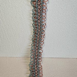 May include: A macrame plant hanger in shades of teal and copper. The handmade hanger features a series of looped knots, creating a decorative, textured design. The hanger is suspended from a white ring and has a tassel at the bottom.