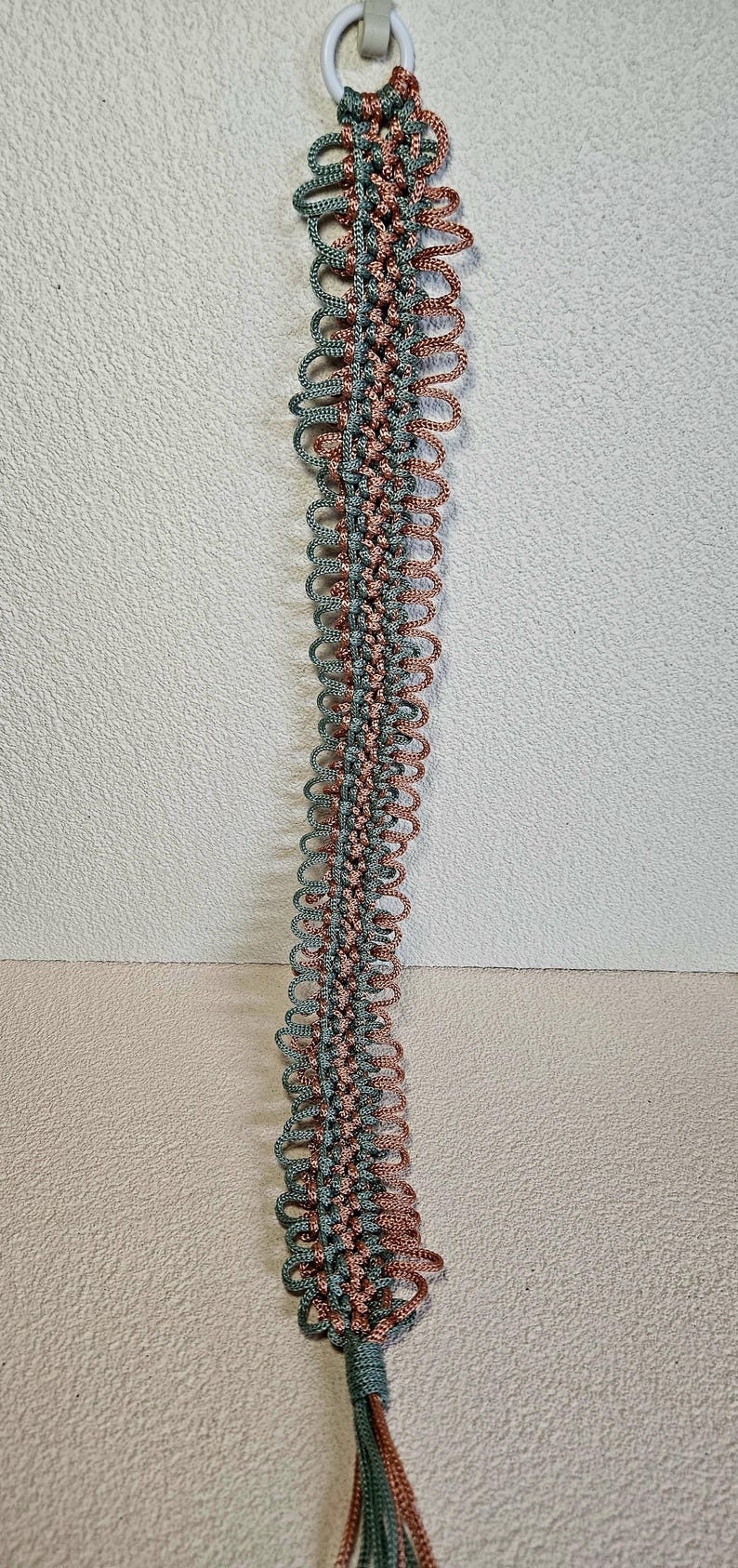 May include: A macrame plant hanger in shades of teal and copper. The handmade hanger features a series of looped knots, creating a decorative, textured design. The hanger is suspended from a white ring and has a tassel at the bottom.