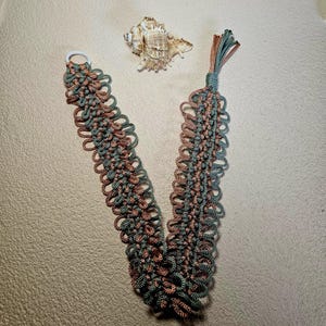 May include: A handmade macrame bookmark in a V-shape, featuring a combination of teal and brown cords. The bookmark has a white ring at one end and a tassel at the other. A seashell rests nearby.