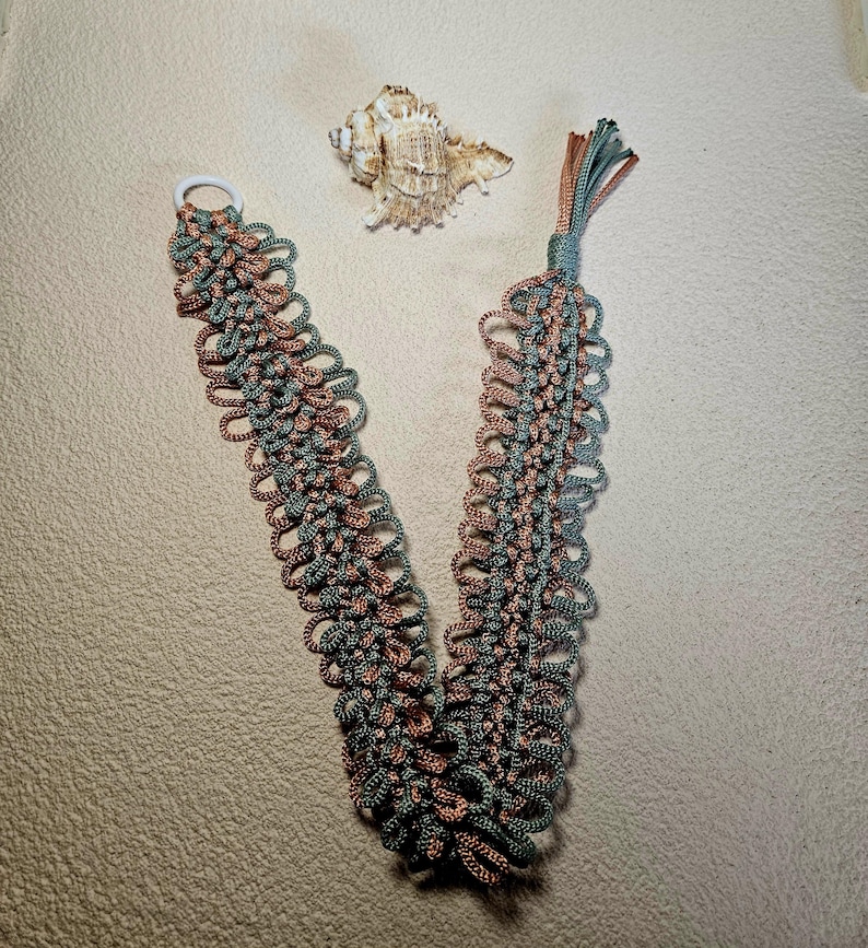 May include: A handmade macrame bookmark in a V-shape, featuring a combination of teal and brown cords. The bookmark has a white ring at one end and a tassel at the other. A seashell rests nearby.