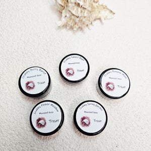 May include: Five small, round containers with black lids and white labels. Each label features text like "Crabby Berry Burst" and "Treat", along with a seashell graphic. A seashell sits above the containers.