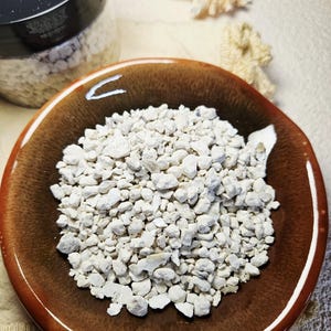May include: A brown ceramic bowl filled with small, irregularly shaped white stones. A jar of similar stones is visible in the background, along with seashells. The jar has a label that reads "Oyster Shells".