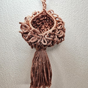 May include: A handmade, copper-colored macrame ornament with a tassel. The ornament features a woven basket shape with intricate looped detailing around the rim. A long tassel hangs from the bottom, and a loop is at the top for hanging.