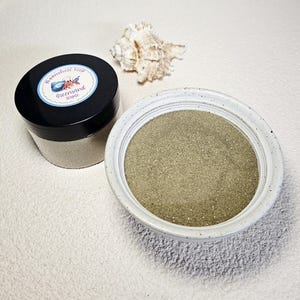 May include: A jar with a black lid and a white ceramic bowl filled with green sand. The jar has a label that reads "MoonShell Reef Greensand staple" with a seashell graphic. A seashell is also present.