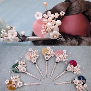May include: A collection of ornate hair accessories, including hairpins and a hair comb. The hairpins feature colorful, circular accents adorned with white flowers and clusters of pearls. The comb has white flowers and pearls. The accessories are set against a blue background.