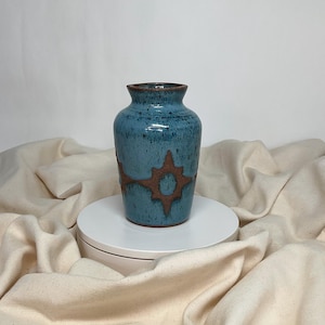 Puede incluir: Un jarrón de cerámica artesanal esmaltado en color turquesa con un diseño de sol marrón. El jarrón tiene un cuerpo redondeado y un cuello estrecho. Este jarrón decorativo es adecuado para exhibir flores o como pieza de arte independiente.