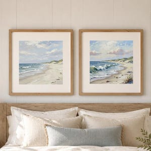 May include: Two square framed wall art prints depicting coastal beach scenes with sand dunes, blue ocean waves, and cloudy skies, displayed in light wood frames above a bed with neutral bedding and a light blue throw blanket.