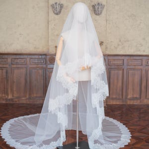 Pode incluir: Um longo véu de noiva branco feito de tule transparente, apresentando uma borda de renda recortada intrincada. O véu está drapeado sobre um manequim, estendendo-se até o chão numa cauda circular com padrões detalhados de renda floral.