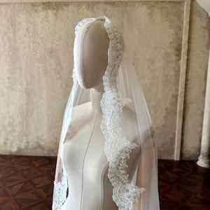 May include: A white wedding veil featuring intricate floral lace trim along the edges. The sheer, lightweight fabric drapes over a mannequin, showcasing the detailed scalloped lace border that frames the face and extends down the length of the veil.