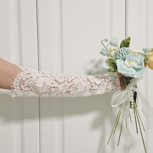 Puede incluir: Mangas de brazo de encaje blanco con un patrón floral. Se sostiene un ramo de flores de ganchillo en colores pastel, incluyendo azul, amarillo y verde. El ramo está atado con una cinta blanca y perlas.