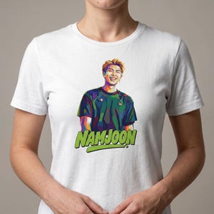 May include: White t-shirt featuring a colorful pop art style portrait of a smiling person. The word "NAMJOON" is printed in green, outlined in black, at the bottom of the shirt.