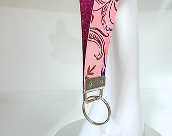 May include: Pink floral print lanyard with a glittery purple backing. The lanyard features a silver key ring and metal hardware. The floral pattern includes blue and brown accents. This accessory is designed to hold keys or an ID badge.