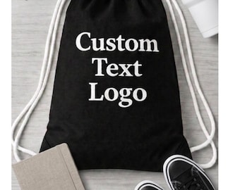 May include: A black drawstring backpack featuring the white text Custom Text Logo printed on the front. The bag has thick white cord straps and is surrounded by sunglasses, a white tumbler, a beige notebook, and a pair of black sneakers.