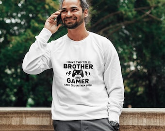 May include: White sweatshirt with the text "I HAVE TWO TITLES BROTHER AND GAMER AND I CRUSH THEM BOTH" in black. The design includes a game controller and lightning bolts. The person in the image is smiling and holding a phone to their ear.
