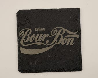 May include: A square, black slate coaster with the words "Enjoy Bour-Bon" etched in a stylized font. The coaster has a slightly rough texture and a natural, dark gray color, suitable for placing under a glass or mug.