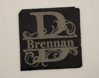 May include: A square, black slate coaster with the name "Brennan" and the letter "D" engraved in a light gray color. The letter "D" has decorative swirls, and the name is centered below.