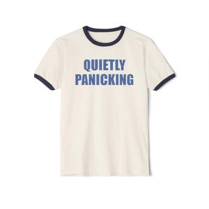 May include: Cream-coloured ringer t-shirt with navy blue trim around the collar and sleeves. The words "QUIETLY PANICKING" are printed in blue block letters across the front. A casual, everyday wear tee.