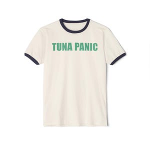 May include: Cream-colored ringer t-shirt with the text "TUNA PANIC" in green block letters. The shirt has navy blue trim around the collar and sleeves. The t-shirt is made of a soft, comfortable material.