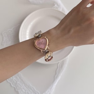 K&ouml;nnte beinhalten: Eine herzf&ouml;rmige Armbanduhr mit rosa Zifferblatt und goldfarbenen Metallakzenten. Die Uhr hat dekorative Blumen- und Edelsteindetails am Armband. Die Uhr befindet sich an einem Handgelenk.