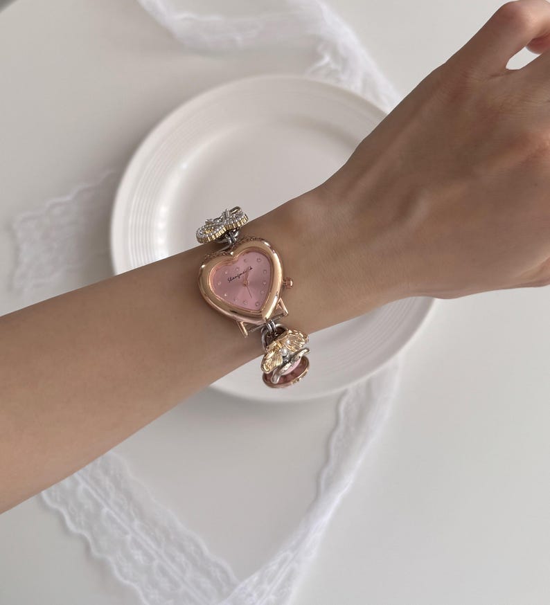K&ouml;nnte beinhalten: Eine herzf&ouml;rmige Armbanduhr mit rosa Zifferblatt und goldfarbenen Metallakzenten. Die Uhr hat dekorative Blumen- und Edelsteindetails am Armband. Die Uhr befindet sich an einem Handgelenk.