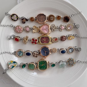 Può includere: Cinque braccialetti decorativi con catene argentate e vari quadranti. I quadranti sono di diverse forme e colori, tra cui rosa, verde e blu. I braccialetti hanno ciondoli decorativi.