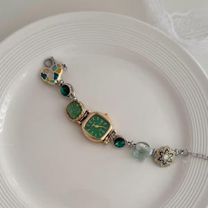 K&ouml;nnte beinhalten: Eine goldfarbene Uhr mit quadratischem gr&uuml;nem Zifferblatt und silberner Kette. Das Uhrenarmband hat gr&uuml;ne Edelsteine, einen herzf&ouml;rmigen Anh&auml;nger und ein Blumenmuster. Die Uhr wird auf einem wei&szlig;en Teller pr&auml;sentiert.