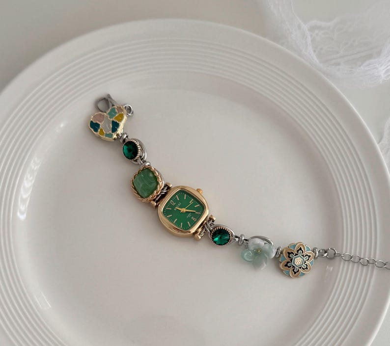 K&ouml;nnte beinhalten: Eine goldfarbene Uhr mit quadratischem gr&uuml;nem Zifferblatt und silberner Kette. Das Uhrenarmband hat gr&uuml;ne Edelsteine, einen herzf&ouml;rmigen Anh&auml;nger und ein Blumenmuster. Die Uhr wird auf einem wei&szlig;en Teller pr&auml;sentiert.