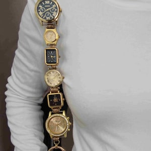 May include: A bag strap constructed from a vertical series of gold-toned wristwatches of varying shapes and sizes, including round and rectangular faces with black and gold dials, connected by metal links and rings, resting on a white long-sleeve shirt.