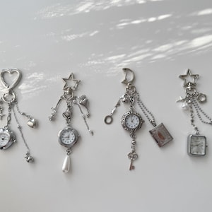 May include: Four silver-toned keychains with miniature watches and decorative charms. Each keychain features a different top shape: a heart, a star, a crescent moon, and another star. Charms include stars, hearts, and a small book.