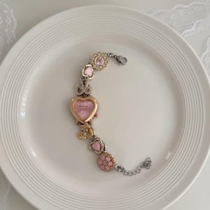 K&ouml;nnte beinhalten: Eine dekorative Armbanduhr mit einem rosa herzf&ouml;rmigen Zifferblatt und einer goldfarbenen L&uuml;nette. Das Armband verf&uuml;gt &uuml;ber rosa- und goldfarbene Charms, darunter Herzen, Blumen und eine Schleife. Die Uhr liegt auf einem wei&szlig;en Teller.