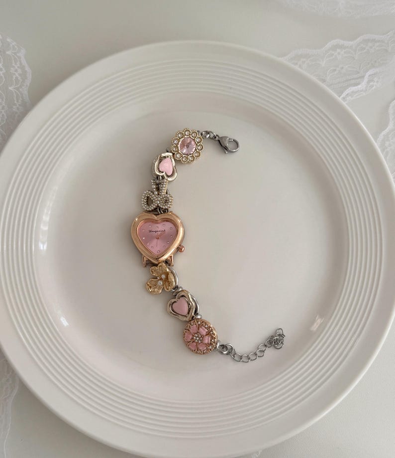 K&ouml;nnte beinhalten: Eine dekorative Armbanduhr mit einem rosa herzf&ouml;rmigen Zifferblatt und einer goldfarbenen L&uuml;nette. Das Armband verf&uuml;gt &uuml;ber rosa- und goldfarbene Charms, darunter Herzen, Blumen und eine Schleife. Die Uhr liegt auf einem wei&szlig;en Teller.