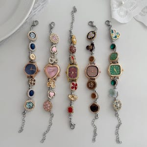 K&ouml;nnte beinhalten: F&uuml;nf dekorative Armbanduhren mit verschiedenfarbigen Zifferbl&auml;ttern und dekorativen Anh&auml;ngern. Die Zifferbl&auml;tter sind in Blau-, Rosa- und Gr&uuml;nt&ouml;nen gehalten. Zu den Anh&auml;ngern geh&ouml;ren Herzen, Blumen und Edelsteine. Die Uhren haben silberfarbene Kettenarmb&auml;nder.