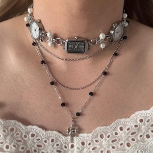 Könnte beinhalten: Mehrschichtige Halskette mit einem Choker mit kleinen Uhren, Kunstperlen und einer silbernen Kette. Die Halskette hat eine zweite Schicht mit schwarzen Perlen und eine dritte Schicht mit einem Kreuzanhänger.